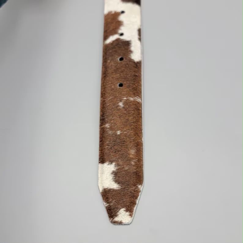 A close-up of a brown and white cowhide belt tip with multiple holes, shown on a neutral light gray background.