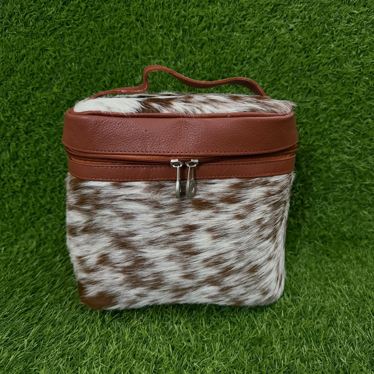 A small rectangular zippered case with a brown faux-leather top and handle, combined with a brown-and-white cowhide lower section, shown on a green grass-like background.