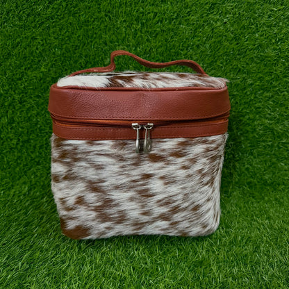 A small rectangular zippered case with a brown faux-leather top and handle, combined with a brown-and-white cowhide lower section, shown on a green grass-like background.