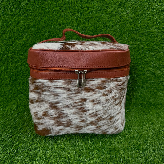 A small rectangular zippered case with a brown faux-leather top and handle, combined with a brown-and-white cowhide lower section, shown on a green grass-like background.