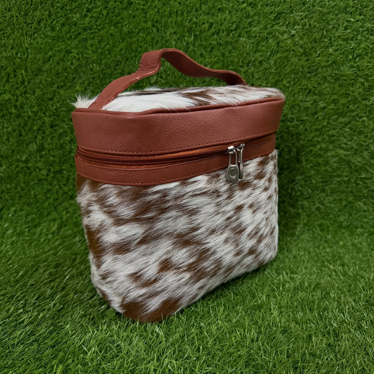 A small zippered cosmetic or travel pouch with a brown and white cowhide pattern body, a smooth reddish-brown top panel, and a top handle, photographed on artificial green grass.