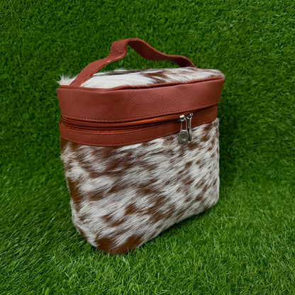 A small zippered cosmetic or travel pouch with a brown and white cowhide pattern body, a smooth reddish-brown top panel, and a top handle, photographed on artificial green grass.