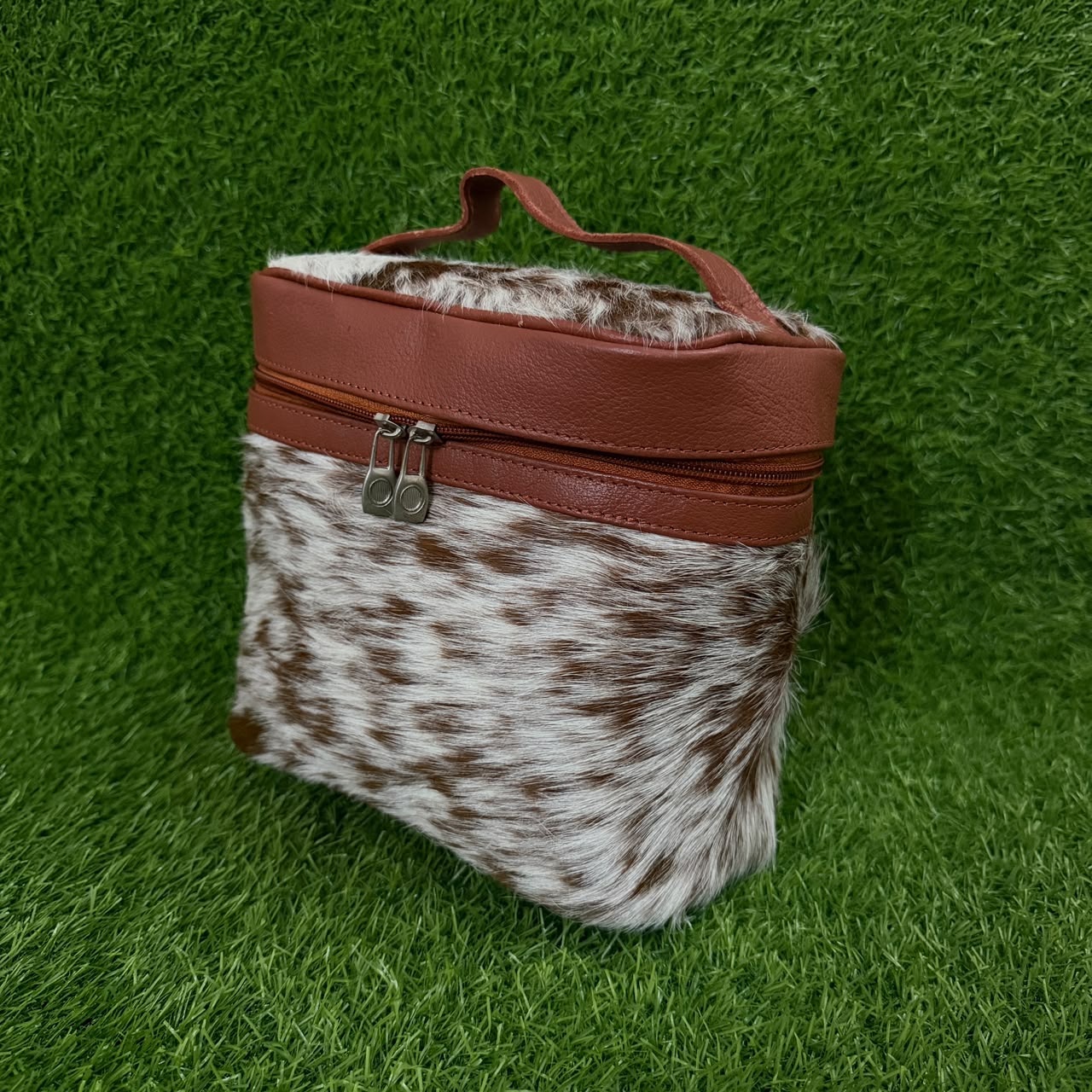 A small zippered bag with a brown and white cowhide-patterned body and reddish-brown faux-leather trim, including a top handle and zipper pull, photographed on a green grass-like surface.