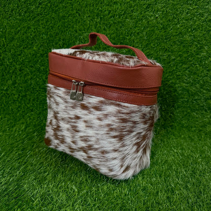 A small zippered bag with a brown and white cowhide-patterned body and reddish-brown faux-leather trim, including a top handle and zipper pull, photographed on a green grass-like surface.