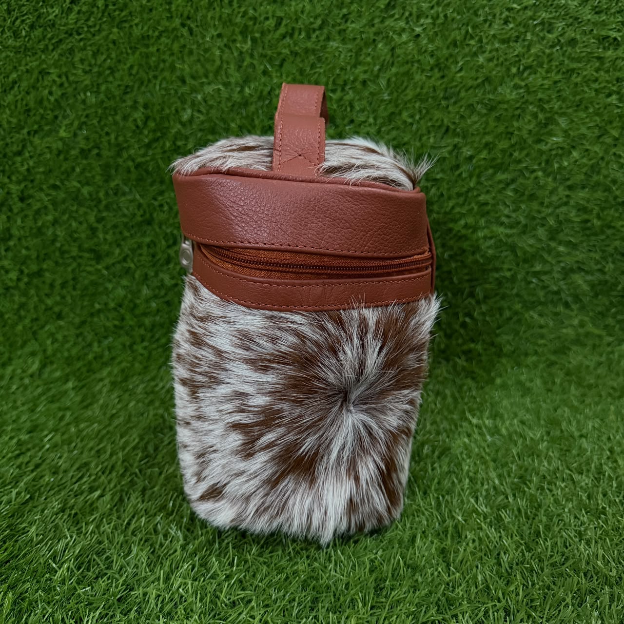 A small cylindrical bag with a brown and white mottled faux fur body, a brown faux leather top band, and a top handle, photographed standing upright on green grass.