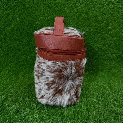 A small cylindrical bag with a brown and white mottled faux fur body, a brown faux leather top band, and a top handle, photographed standing upright on green grass.