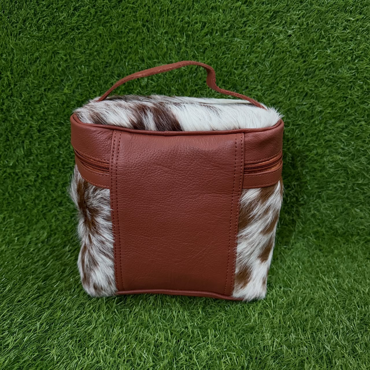 A small rectangular bag with a reddish-brown faux-leather front and handle, combined with brown-and-white cowhide panels on the sides, photographed on a green grass-like surface.