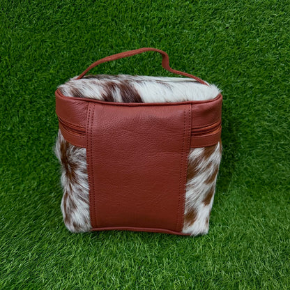 A small rectangular bag with a reddish-brown faux-leather front and handle, combined with brown-and-white cowhide panels on the sides, photographed on a green grass-like surface.