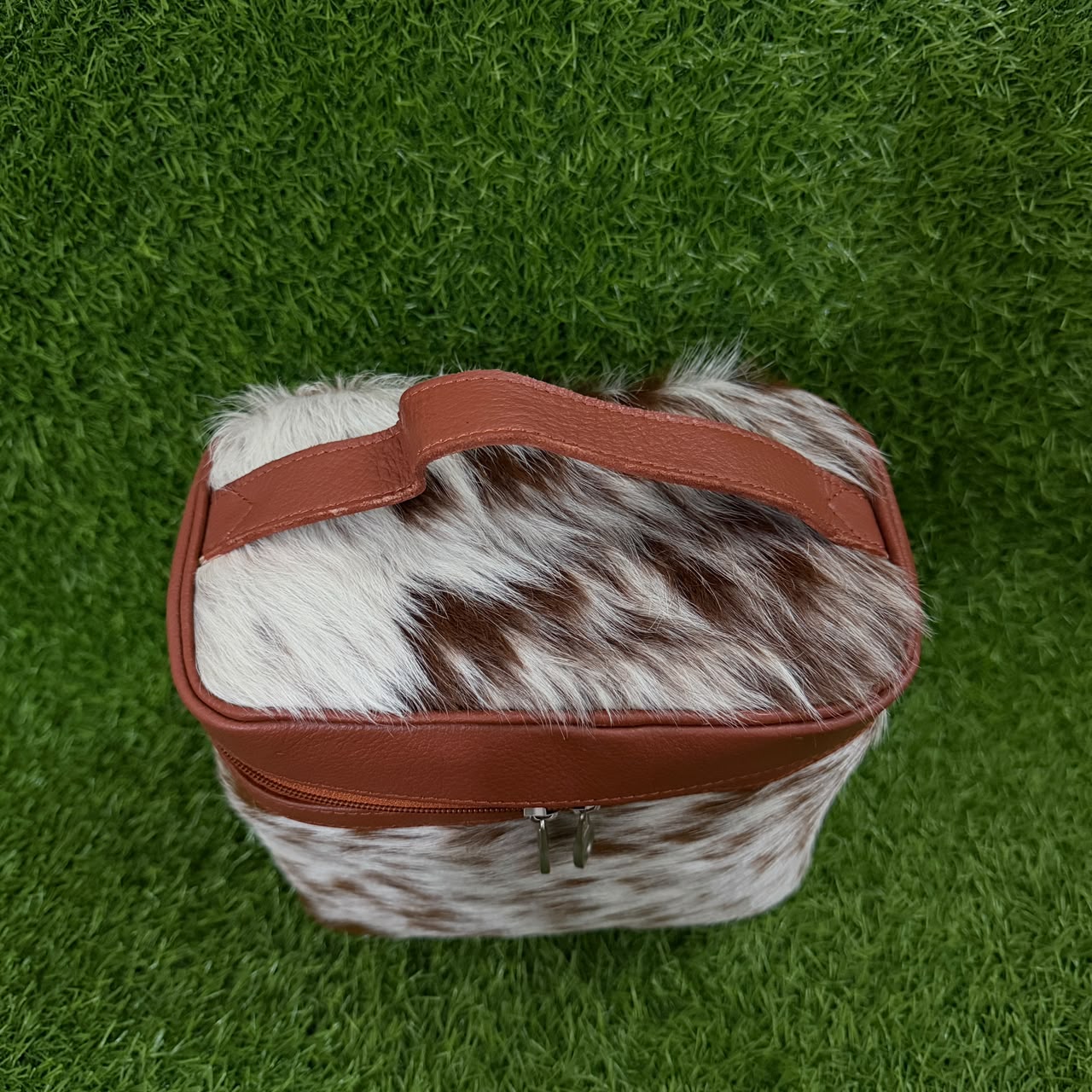 A rectangular zippered pouch or small bag with a brown and white cowhide patterned body and reddish-brown faux-leather trim and handle, photographed from above on a green grass-like surface.