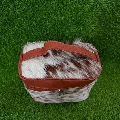 A rectangular zippered pouch or small bag with a brown and white cowhide patterned body and reddish-brown faux-leather trim and handle, photographed from above on a green grass-like surface.