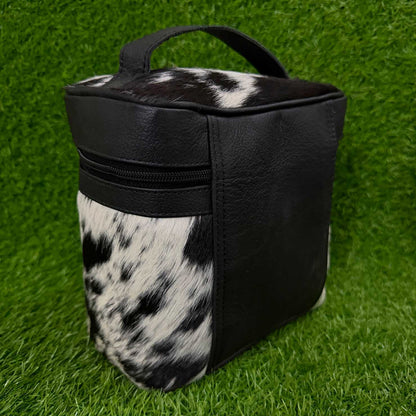 A black and black-and-white cowhide-patterned soft-sided bag with a top handle, standing on artificial green grass.