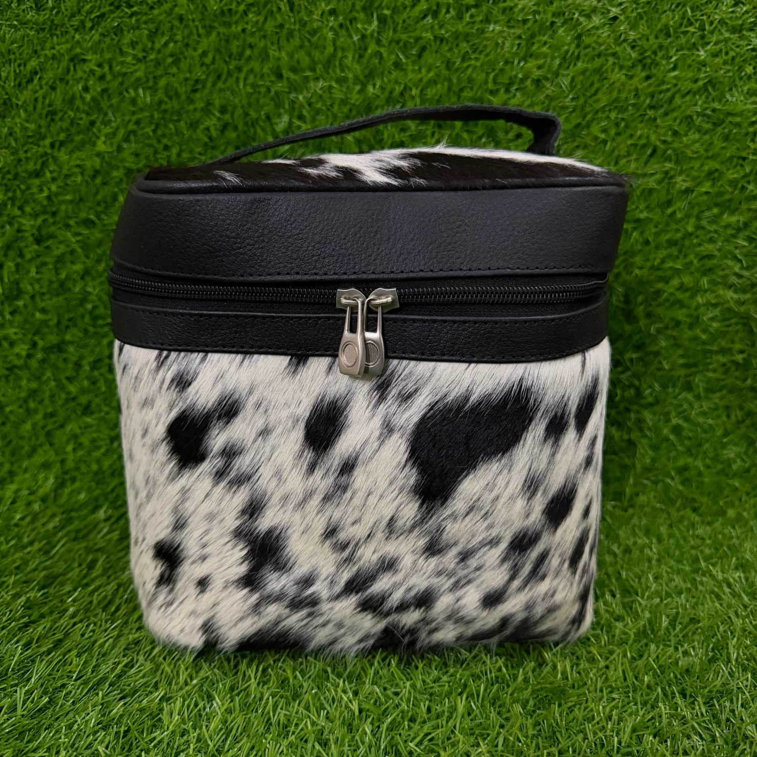 A rectangular zippered cosmetic bag with a black faux-leather top and handle, and a black-and-white cowhide patterned body, shown on a green grass-like background.