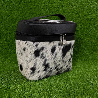 A  zippered cosmetic bag with a black faux-leather top and handle, and a black-and-white cowhide-patterned body, shown on a green grass background.