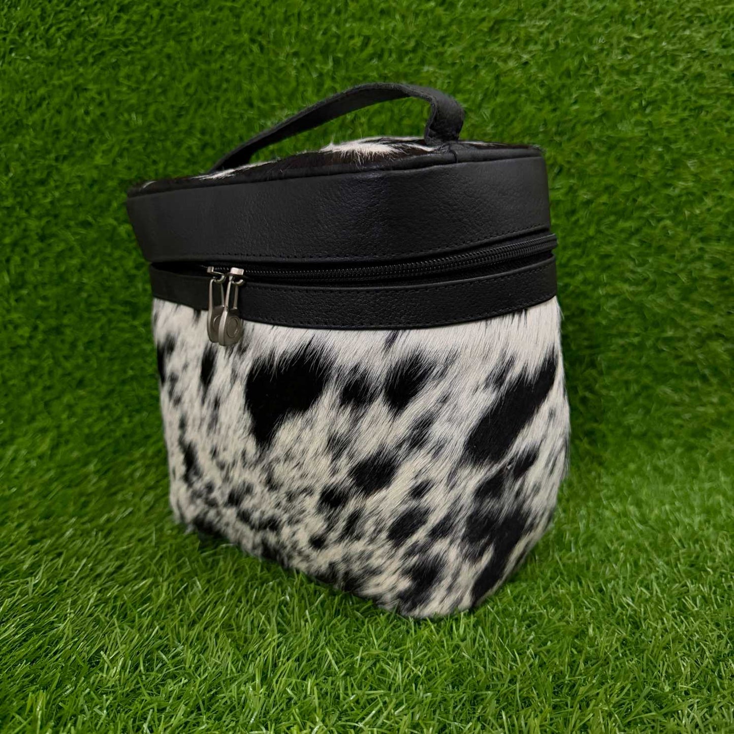 A cylindrical zippered pouch or case with a black faux-leather top band and handle, and a black-and-white cowhide-patterned body, photographed on bright green artificial grass.