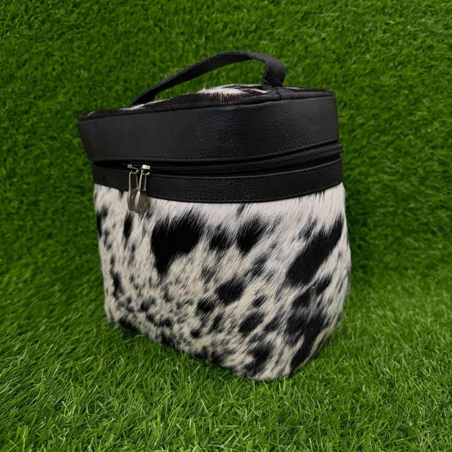 A cylindrical zippered pouch or case with a black faux-leather top band and handle, and a black-and-white cowhide-patterned body, photographed on bright green artificial grass.
