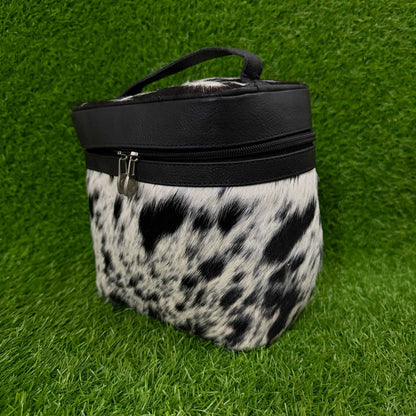 A cylindrical zippered pouch or case with a black faux-leather top band and handle, and a black-and-white cowhide-patterned body, photographed on bright green artificial grass.