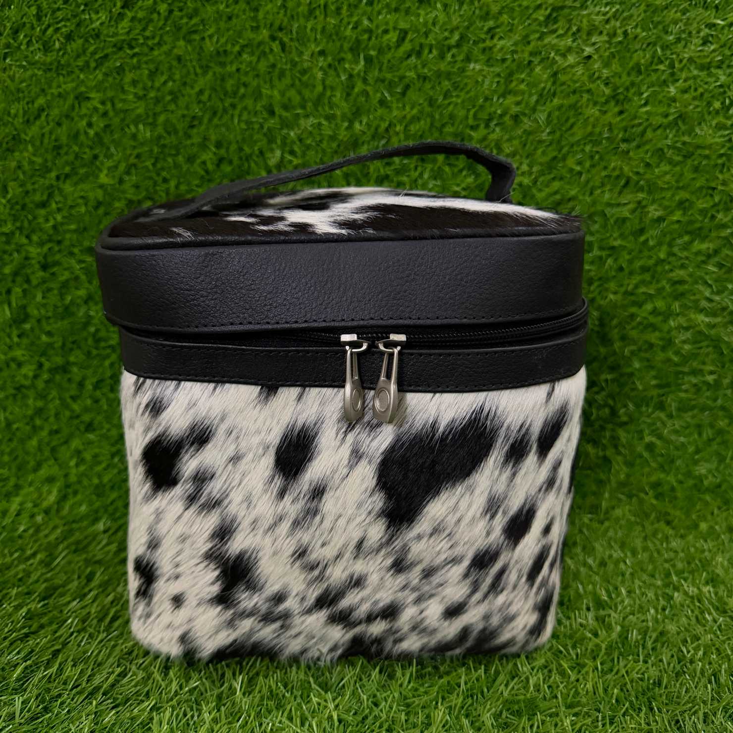 A cylindrical zippered cosmetic bag with a black faux-leather top band and handle, and a black-and-white cowhide-patterned body, shown on a green grass-like surface.