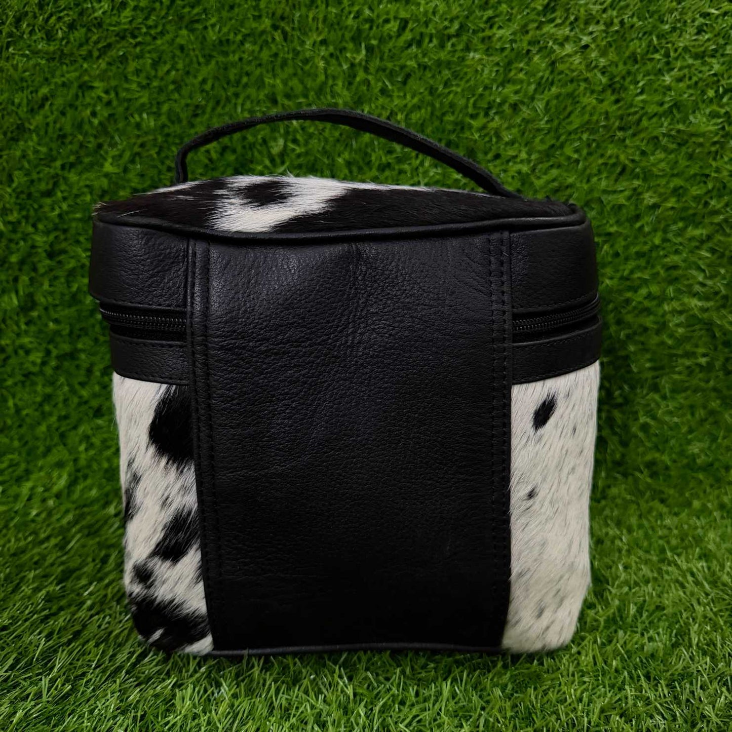 A small black and white cowhide zippered pouch with a black faux-leather front panel and top handle, photographed on a green grass-like surface.