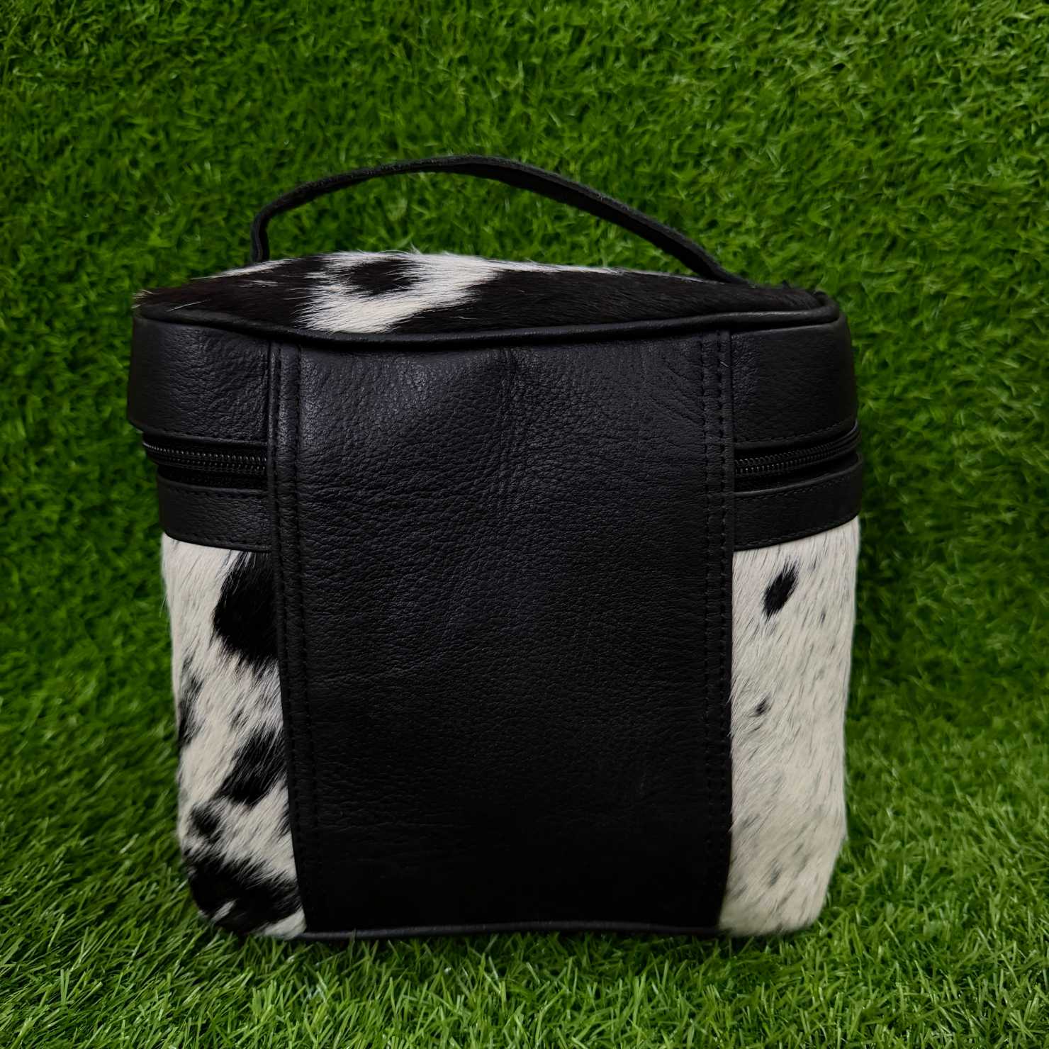 A small black and white cowhide zippered pouch with a black faux-leather front panel and top handle, photographed on a green grass-like surface.