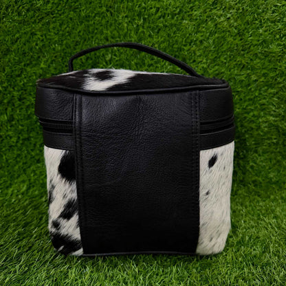 A small black and white cowhide zippered pouch with a black faux-leather front panel and top handle, photographed on a green grass-like surface.