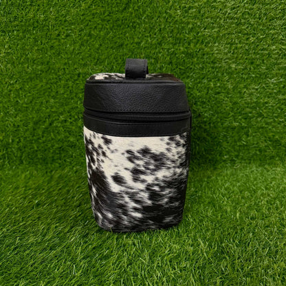 A small cylindrical bag or container with a black faux-leather top and handle, featuring a black and white cowhide pattern body, set against a green artificial grass background.