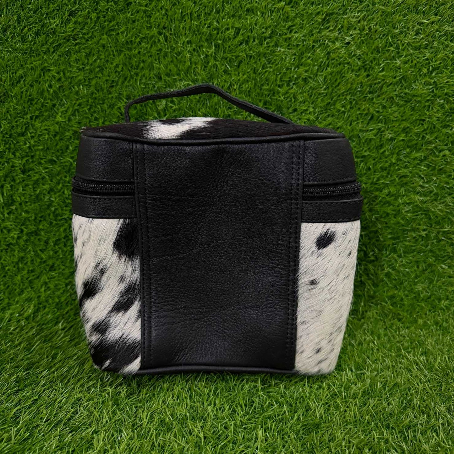 A small black and black-and-white cowhide pouch with a top handle, photographed on a green grass-like surface.