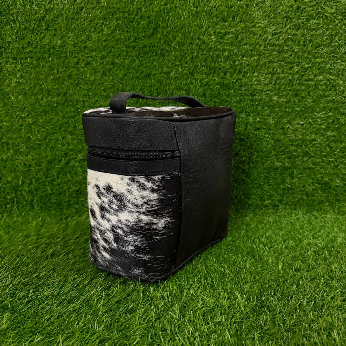 A black and black-and-white cowhide-patterned soft-sided bag with a top handle, standing on artificial green grass.