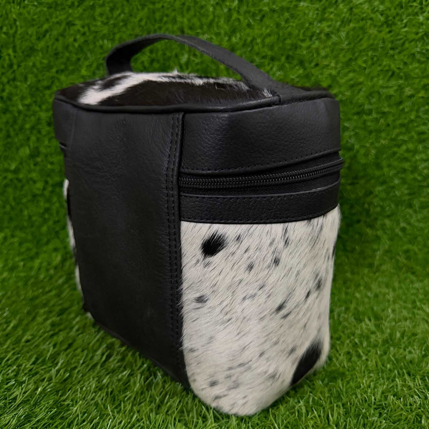 A small black and black-and-white cowhide zippered pouch or organizer bag with a top handle, photographed on artificial green grass.