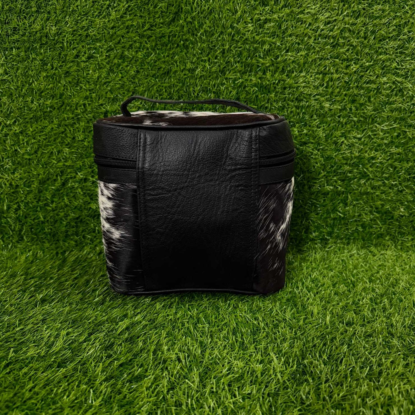 A black leather zippered cosmetic bag with black and white cowhide side panels and a top handle, set against a green grass background.