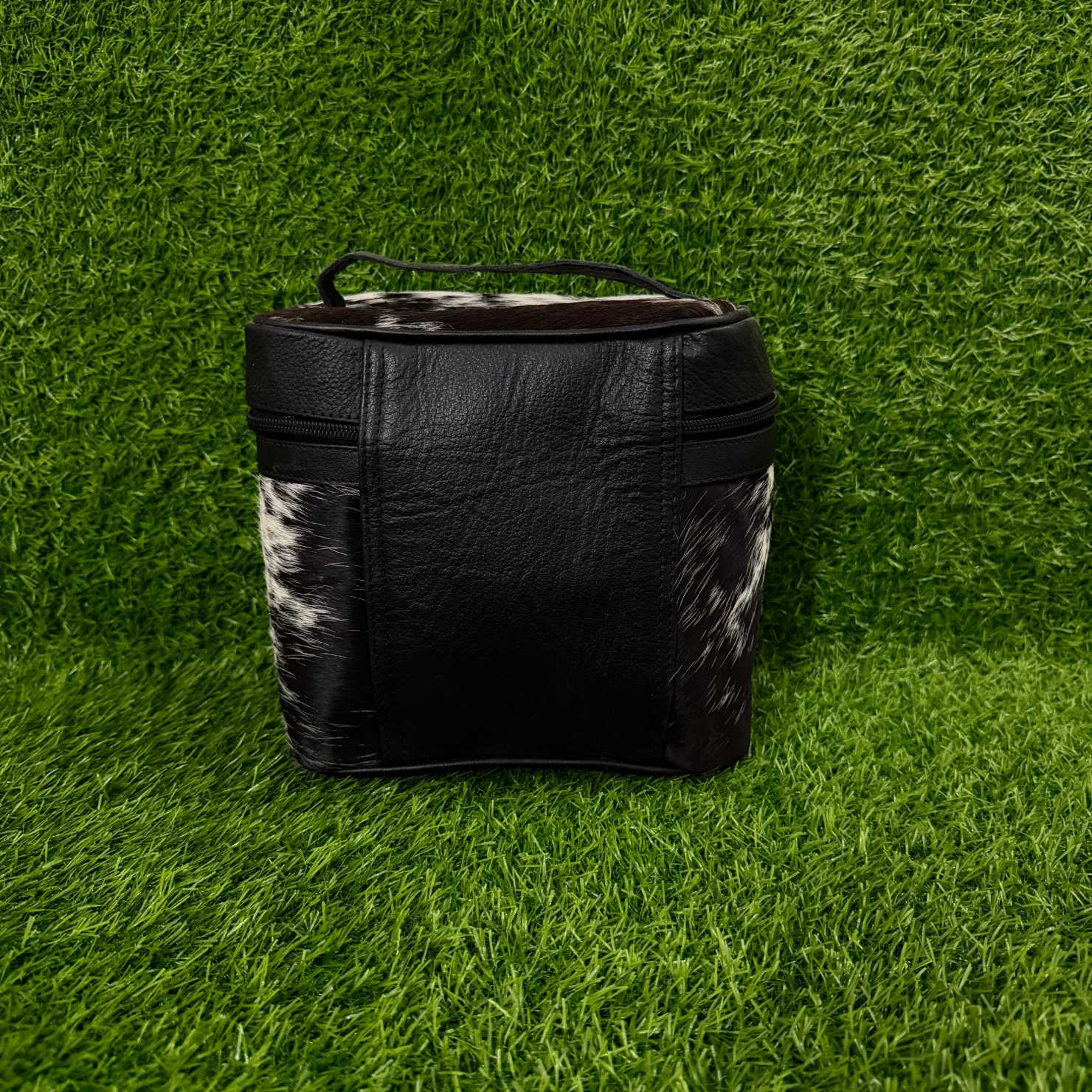 A black leather zippered cosmetic bag with black and white cowhide side panels and a top handle, set against a green grass background.