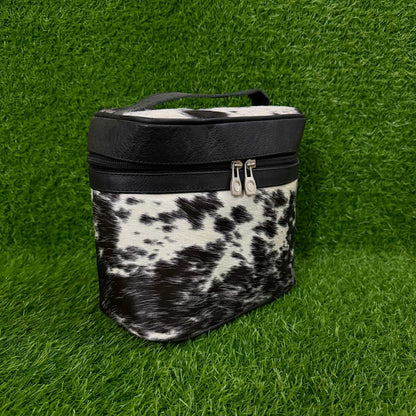 A black and white cow-print cosmetic bag with black faux-leather trim and a top handle, set against a green grass background.