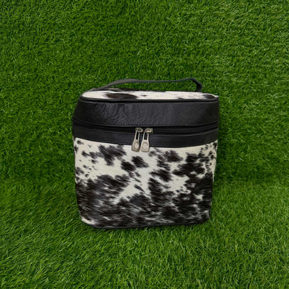 A small zippered cosmetic or storage bag with a black faux-leather top band and handle, and a black-and-white cowhide-pattern lower body, photographed on bright green artificial grass.