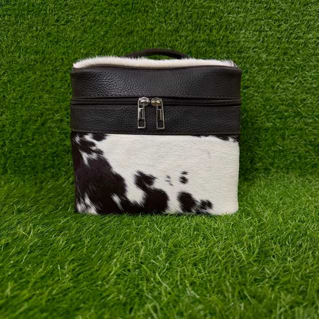 A rectangular cosmetic bag with a black textured leather top, silver zipper, white faux-fur trim, and a black-and-white cowhide lower section, displayed on a green grass background.