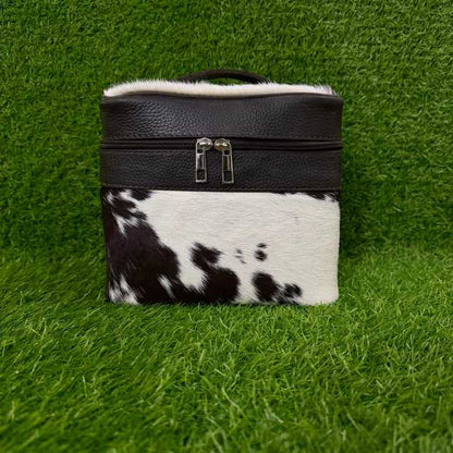 A rectangular cosmetic bag with a black textured leather top, silver zipper, white faux-fur trim, and a black-and-white cowhide lower section, displayed on a green grass background.