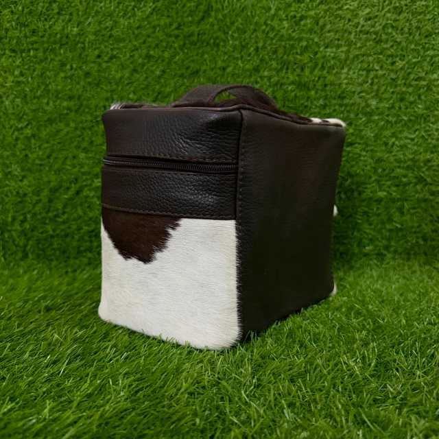 A small rectangular handbag or pouch made of dark brown leather and cowhide, featuring a white and dark brown patch pattern, set against a green grass background.