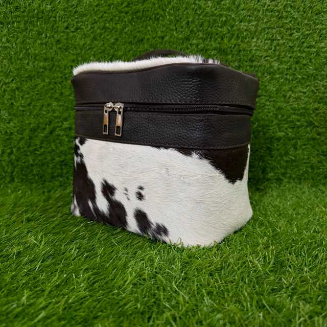A small black and white cowhide zippered pouch or cosmetic case with a black faux-leather top band and silver zipper pulls, photographed on artificial green grass.