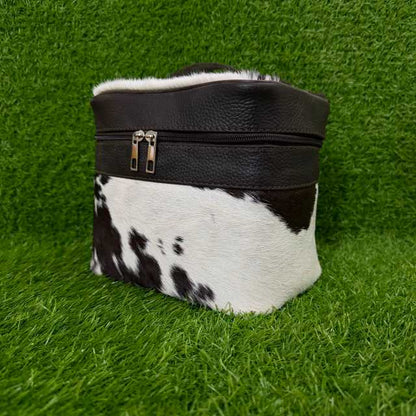 A small black and white cowhide zippered pouch or cosmetic case with a black faux-leather top band and silver zipper pulls, photographed on artificial green grass.