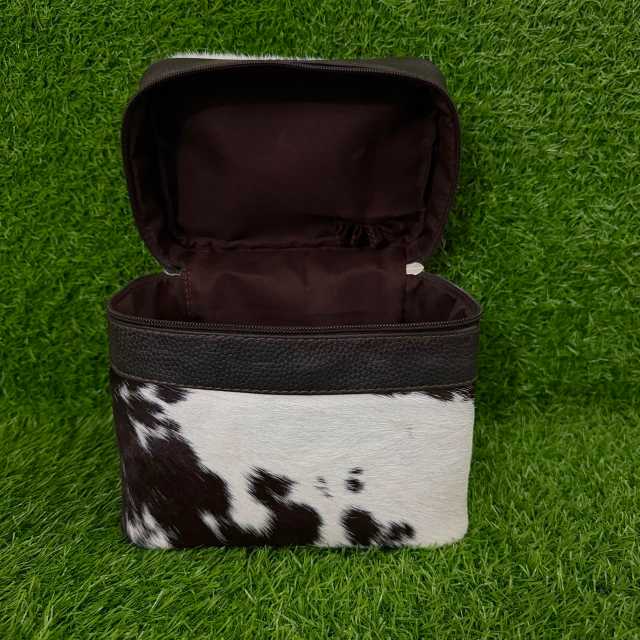 A rectangular zippered cosmetic bag with a black faux-leather top trim and a black-and-white cowhide patterned lower half, shown open on a green grass-like surface.