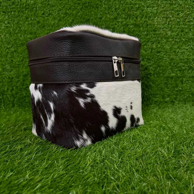A small zippered pouch or cosmetic bag with a black faux-leather top band and a black-and-white cowhide lower section, photographed on artificial green grass.