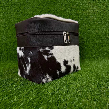 A small zippered pouch or cosmetic bag with a black faux-leather top band and a black-and-white cowhide lower section, photographed on artificial green grass.