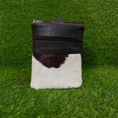 A small leather and cowhide pouch standing upright on green grass. The top half is dark brown pebbled leather with a zipper, and the bottom half is white cowhide with a dark brown patch.