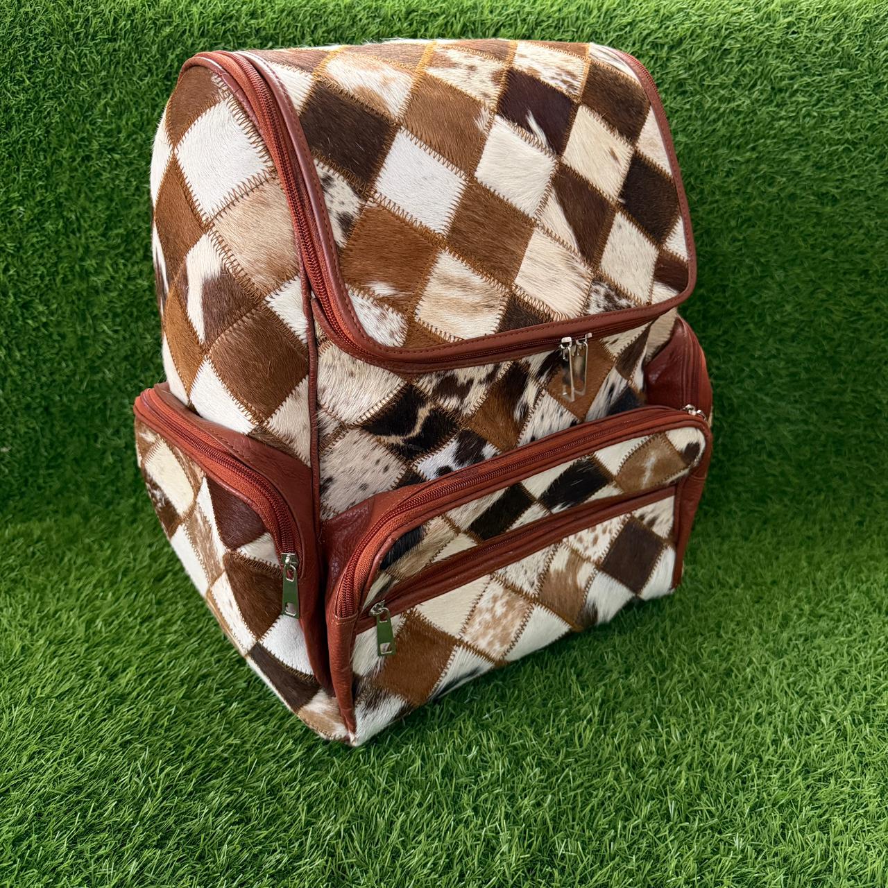 A structured backpack with a brown and cream argyle cowhide pattern and reddish-brown trim, shown on a green artificial grass background.