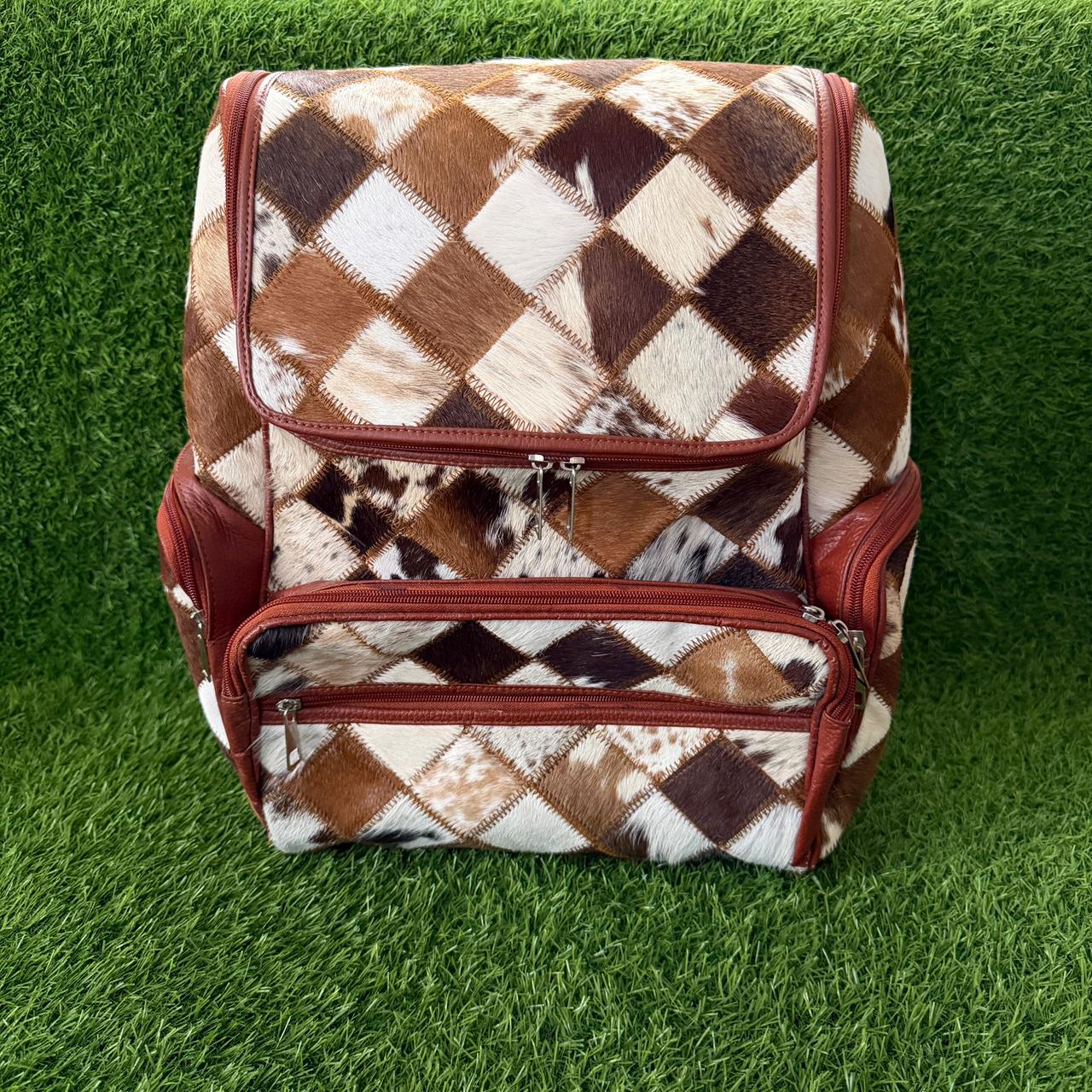 A backpack with a brown, white, and black argyle cowhide pattern and reddish-brown leather trim, shown on a green artificial grass background.