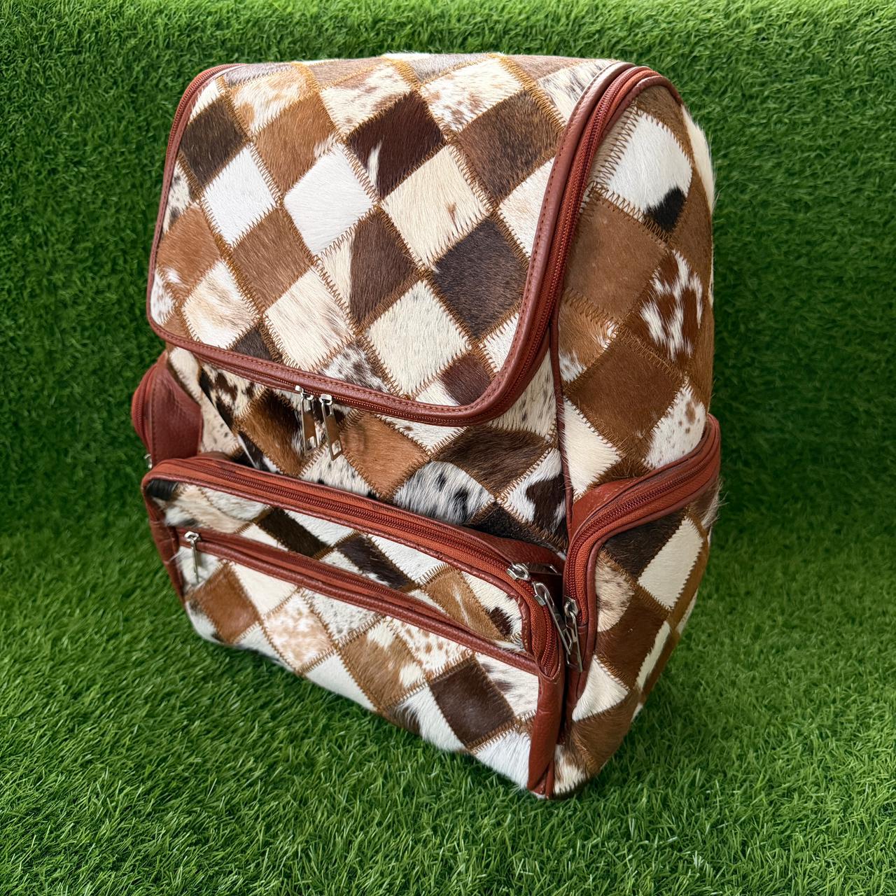 A backpack with a brown, white, and black argyle animal-print pattern and reddish-brown trim, shown on a green artificial grass background.