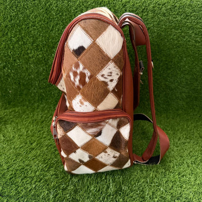 A backpack made of brown and white cowhide with an argyle pattern, featuring brown faux-leather trim and straps, standing upright on a green artificial grass background.