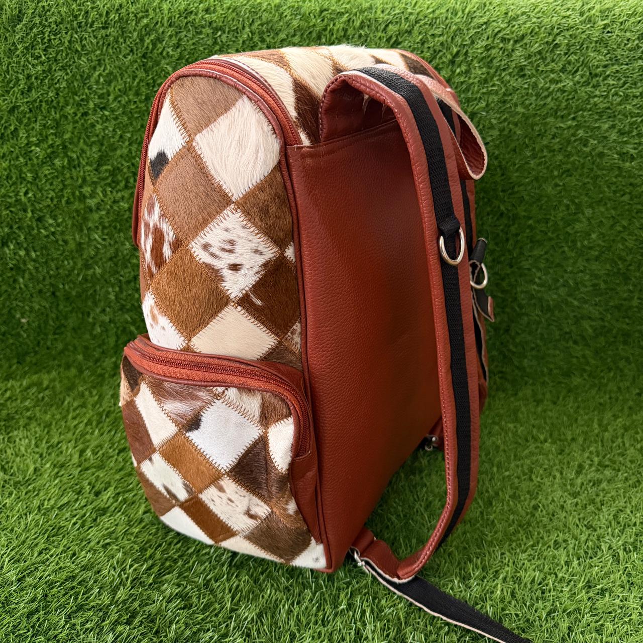 A brown and cream argyle-patterned backpack with smooth brown leather side panels and a black-and-white striped shoulder strap, photographed on artificial green grass.