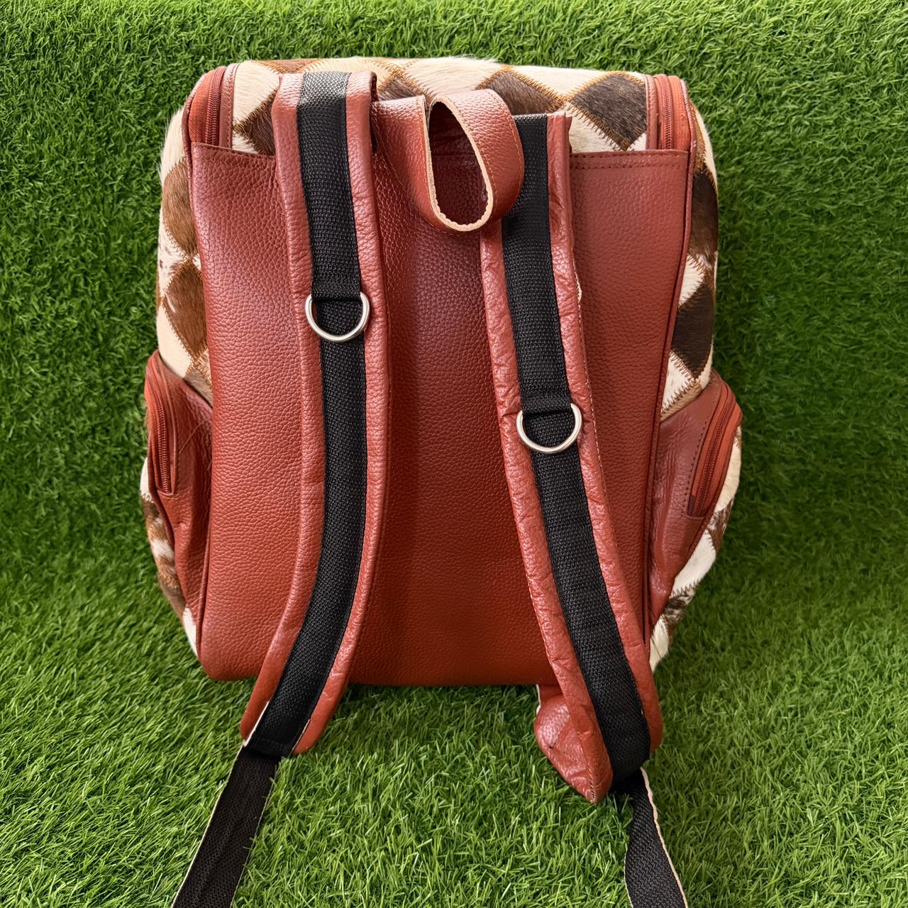 A brown leather backpack shown from the back, featuring textured brown leather panels, black padded shoulder straps, and a brown and beige argyle pattern on the side panels, set against a green artificial grass background.