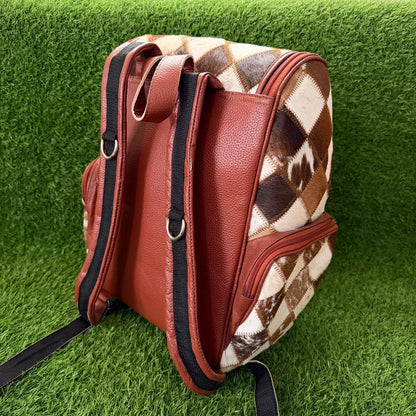 A brown faux-leather backpack with a brown and cream argyle cowhide pattern side panel, shown on a green artificial grass background.
