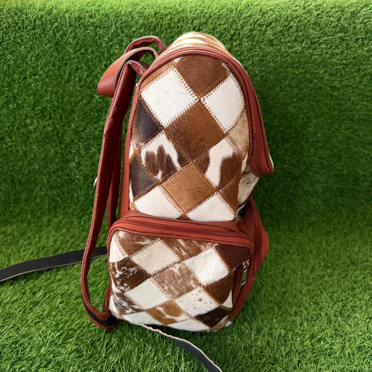 A backpack with a brown and white argyle pattern made from cowhide, featuring reddish-brown leather trim and a shoulder strap, set against a green artificial grass background.