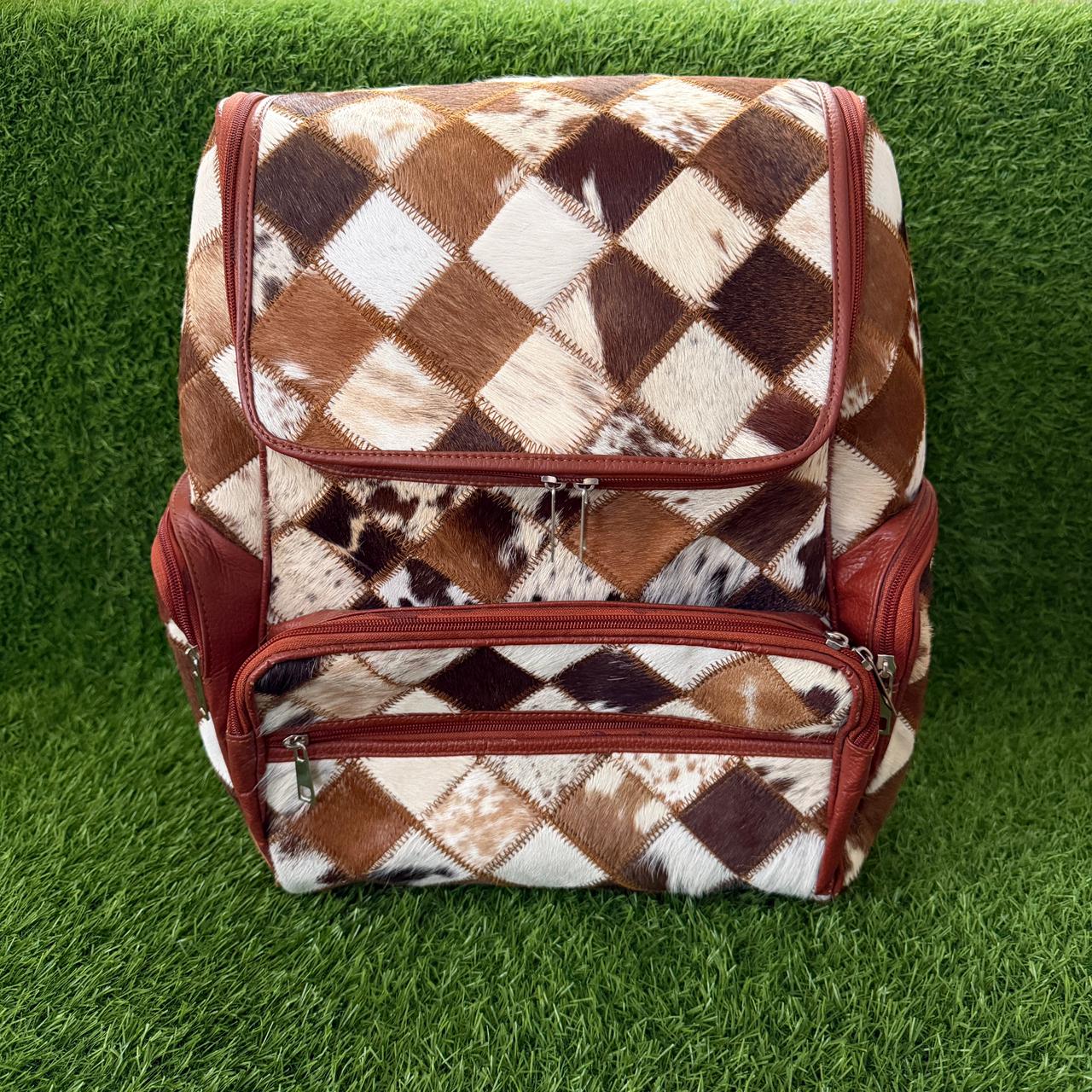 A backpack with a brown, beige, and white cowhide argyle pattern and reddish-brown leather trim, shown on a green grass background.